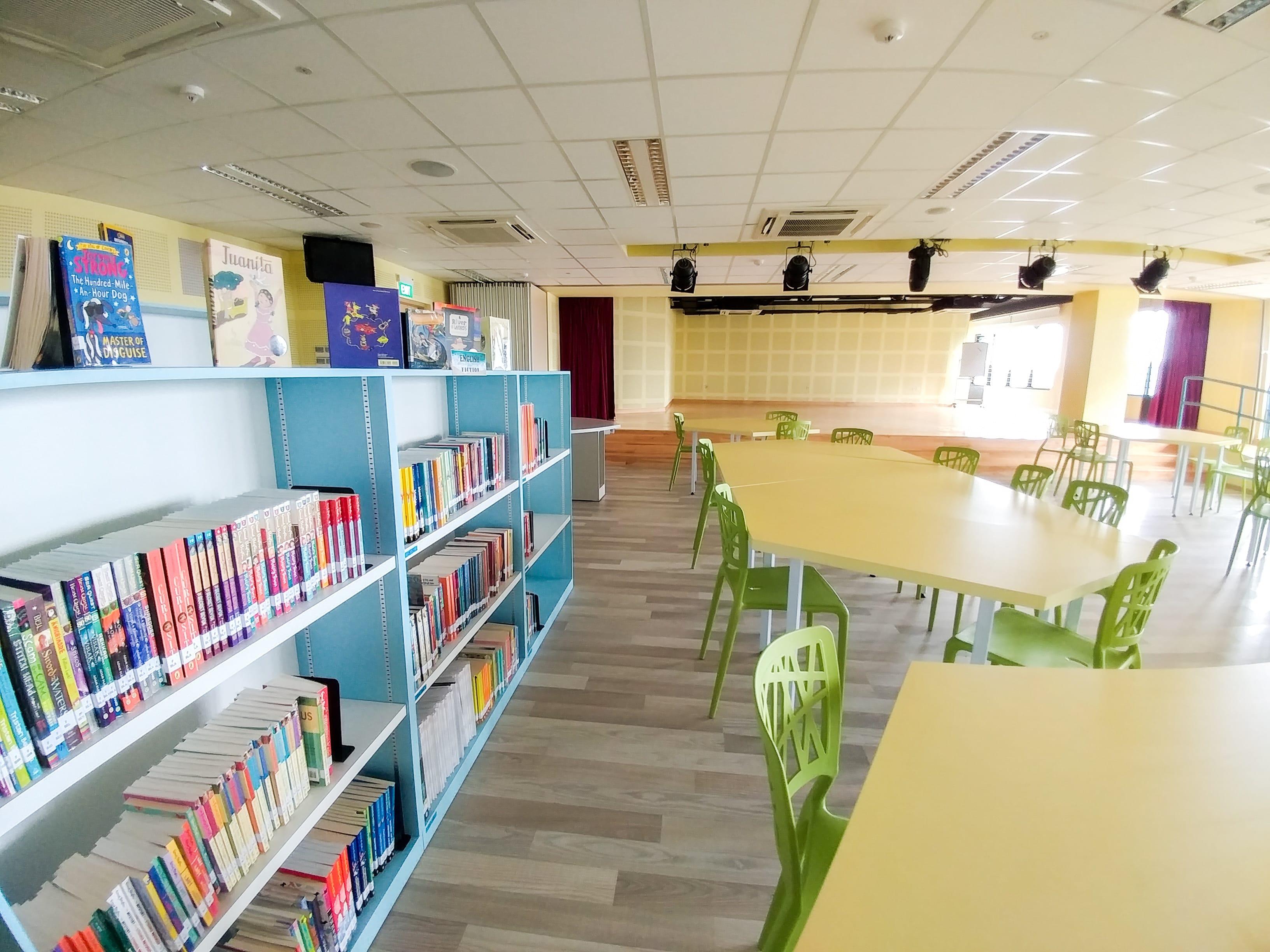 A bright school library with blue bookshelves full of books and yellow tables with green chairs on a wood floor.